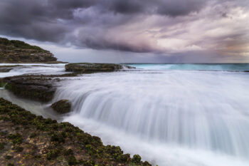 Stormy sunset over Tamarama Beach, Sydney, with waves flowing across the rocky shoreline, fine art coastal photography.