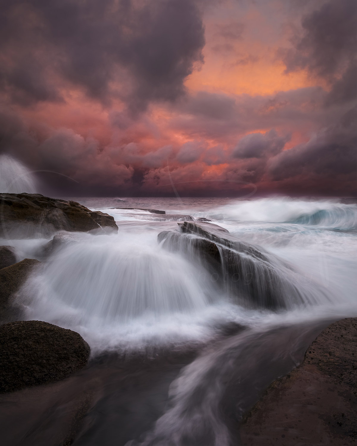 Stormy sunset over Bondi Beach, Sydney, with waves surging across coastal rocks beneath dramatic red and orange clouds.