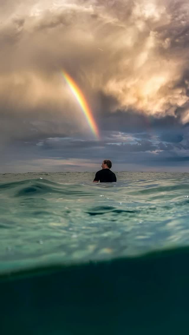 A surfer waiting in the lineup as a rainbow breaks through the storm
