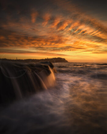 Bondi Beach sunrise with golden clouds and ocean water cascading over a rocky ledge in Sydney, Australia.