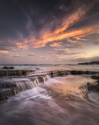 Ocean waves flowing over rocky ledges beneath a colourful sunset sky at Tamarama Beach, Sydney, Australia, coastal photography.