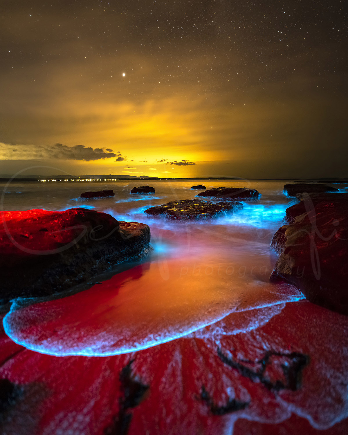 Bioluminescence glowing blue among coastal rocks at night in Jervis Bay, NSW, with stars and golden light on the horizon.