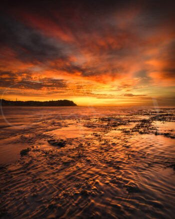Fiery sunrise over Bondi Beach, Sydney, with orange clouds reflected across the ocean and rocky shoreline.