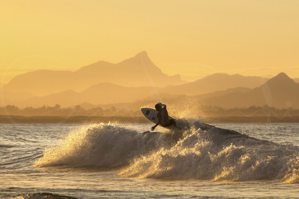 Surfer carving through a wave at Byron Bay at sunset, backlit by golden light with layered mountains creating a dramatic seascape.