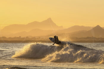 Surfer carving through a wave at Byron Bay at sunset, backlit by golden light with layered mountains creating a dramatic seascape.
