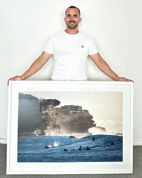 Gergo Rugli holding a large framed award-winning photograph of a bottlenose dolphin leaping among surfers at Bronte Beach, Sydney.
