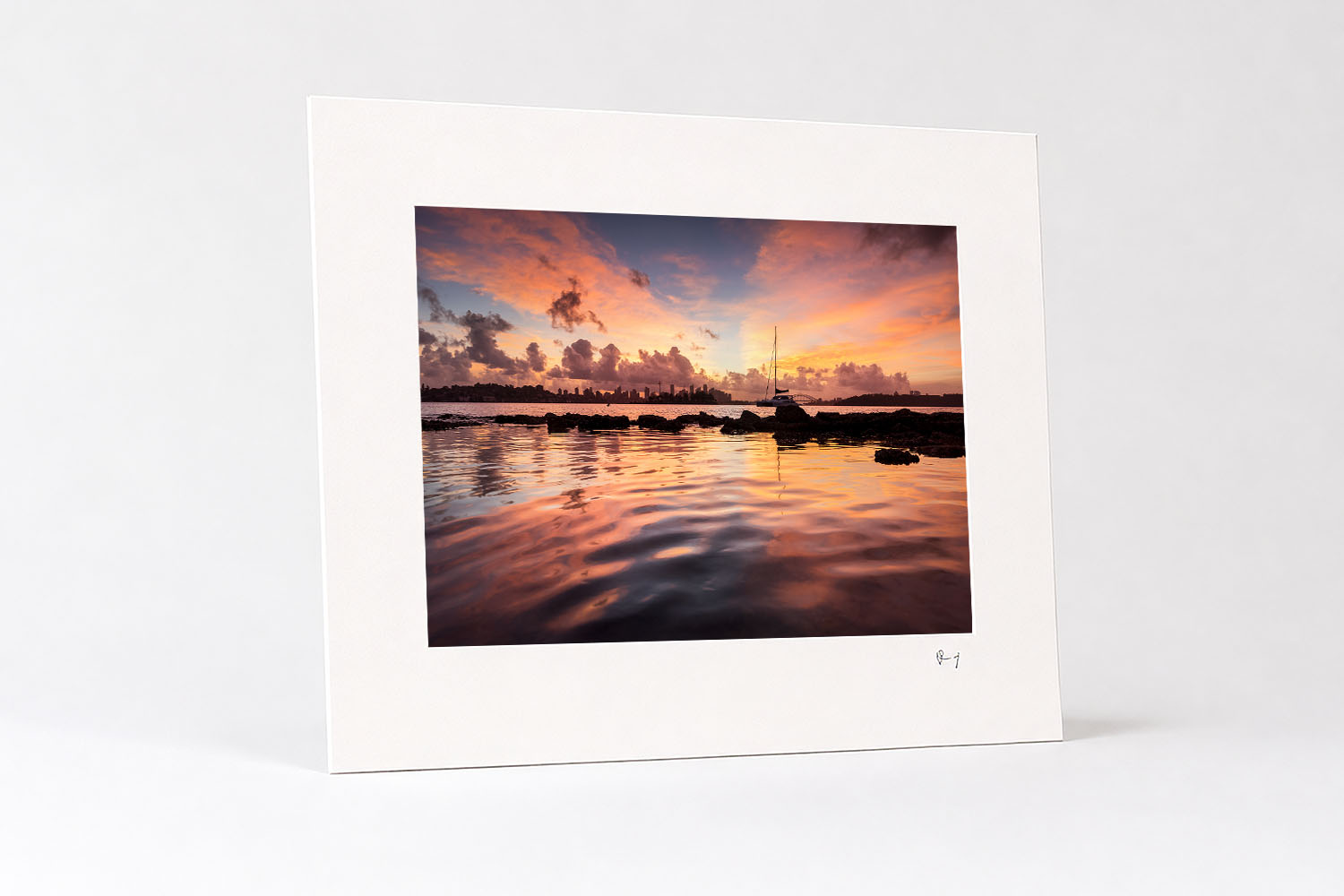 Mounted fine art print of Milk Beach at sunset, with a sailboat beneath vivid pink clouds reflected across the calm harbour.