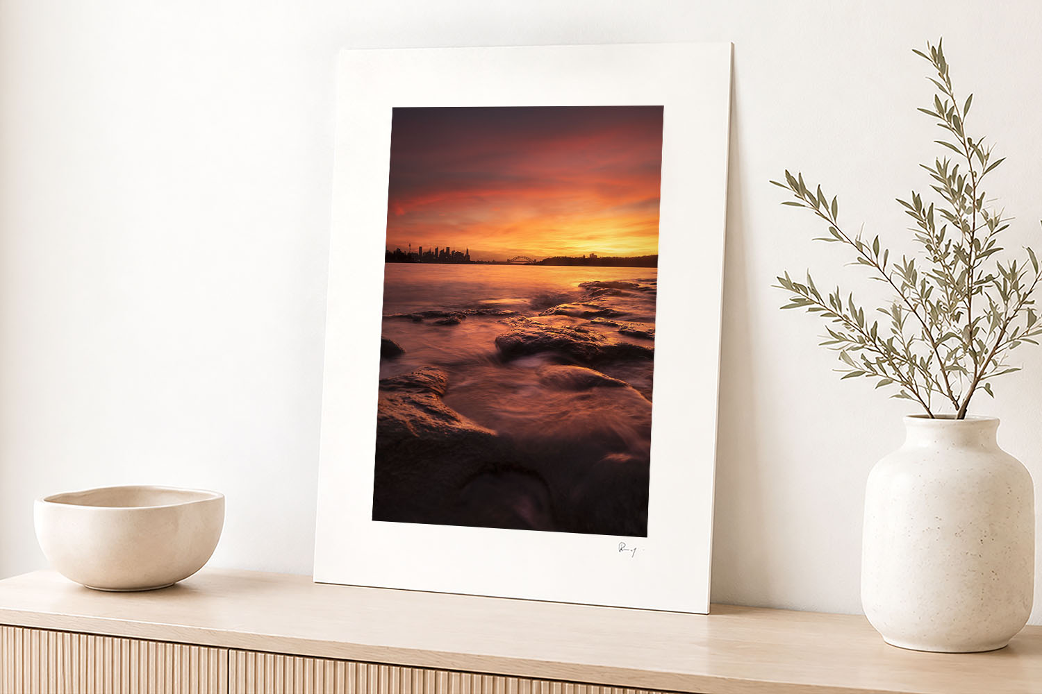 Mounted fine art photograph of a fiery Milk Beach sunset over Sydney Harbour, displayed in a bright contemporary coastal interior.
