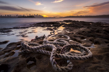 Frayed rope coiled on sandstone at Milk Beach, Sydney Harbour, with sunset reflections and the city skyline in the distance.